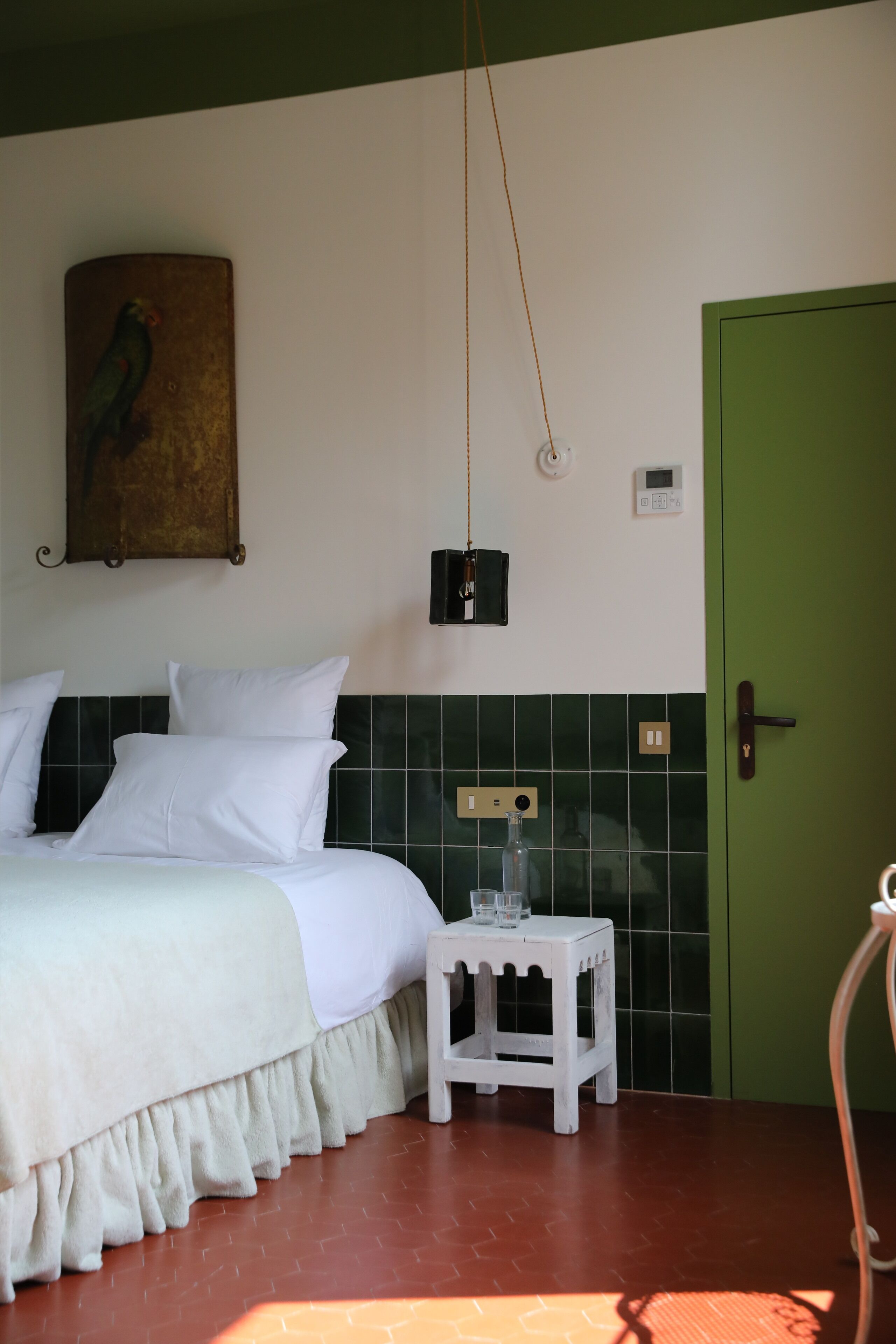 A bedroom with a bed, a small stool, a green door, and a wall-mounted light fixture.