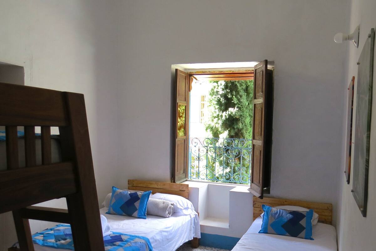 A room with two beds, a window with a view of greenery, and a wooden chair.