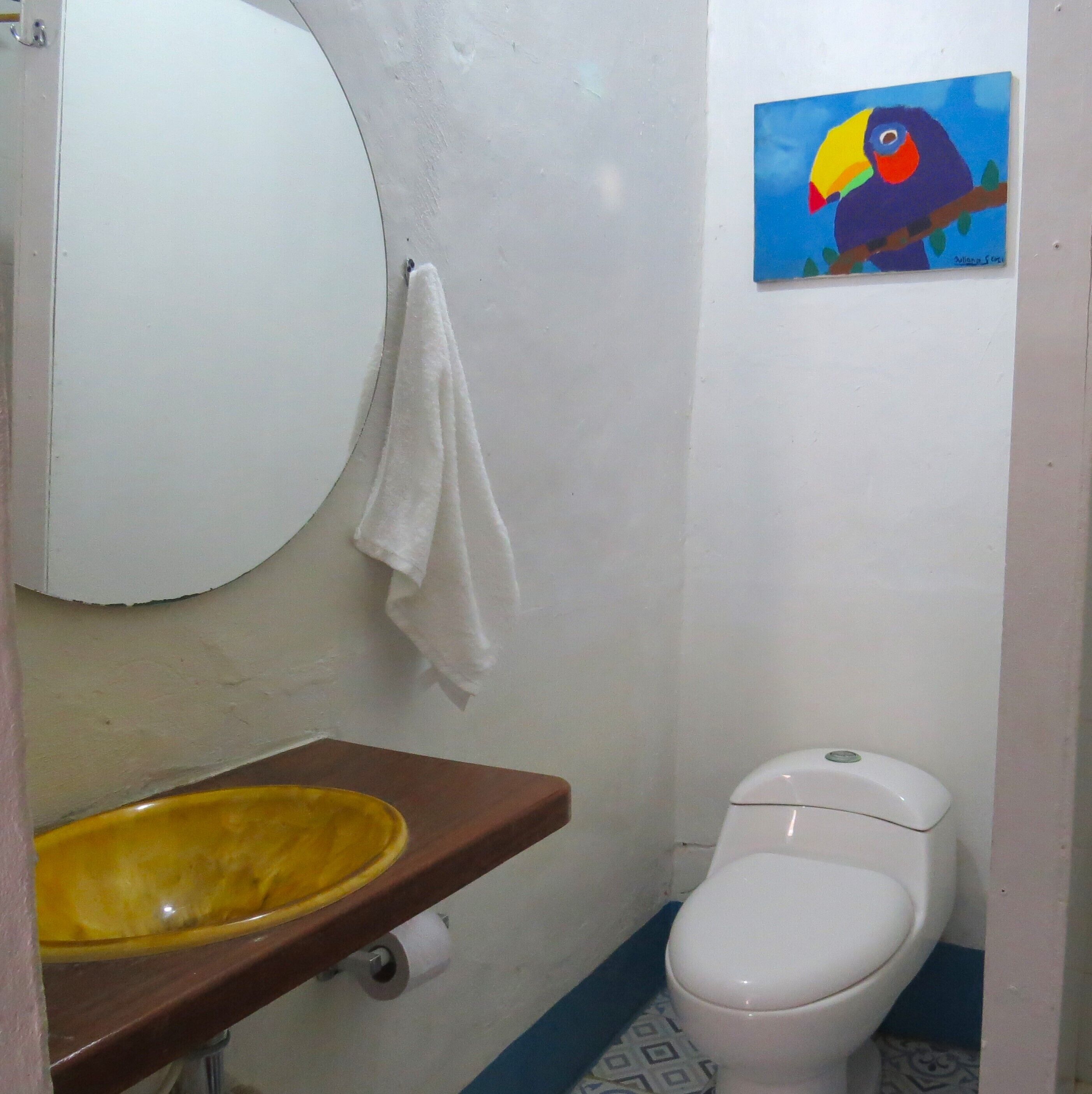 Une salle de bain avec des toilettes blanches, un lavabo jaune et un tableau représentant un oiseau aux couleurs vives accroché au mur.