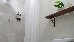 A bathroom with a white toilet, a shower with a curtain, a small shelf with a plant, and a blue mat on the floor. - Casa Fibonacci (Titiribí)