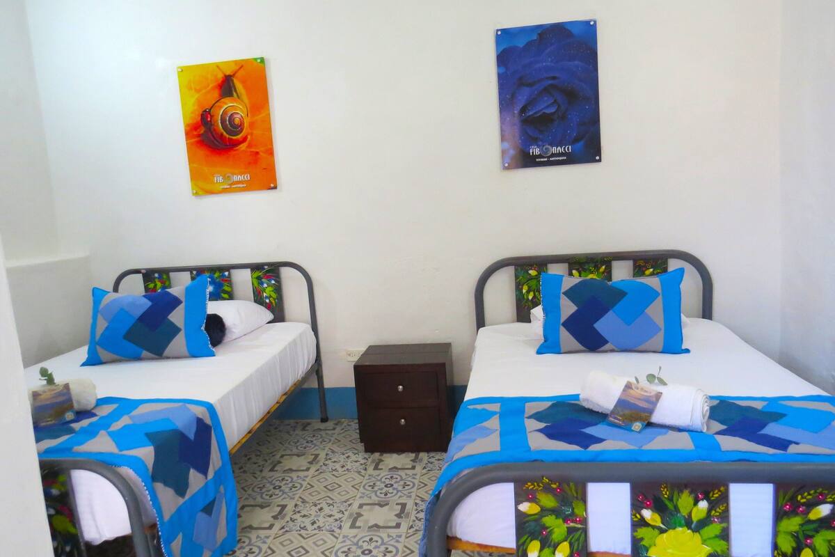 A room with two beds, each with blue and white bedding, a small wooden nightstand, and a wall with two posters.
