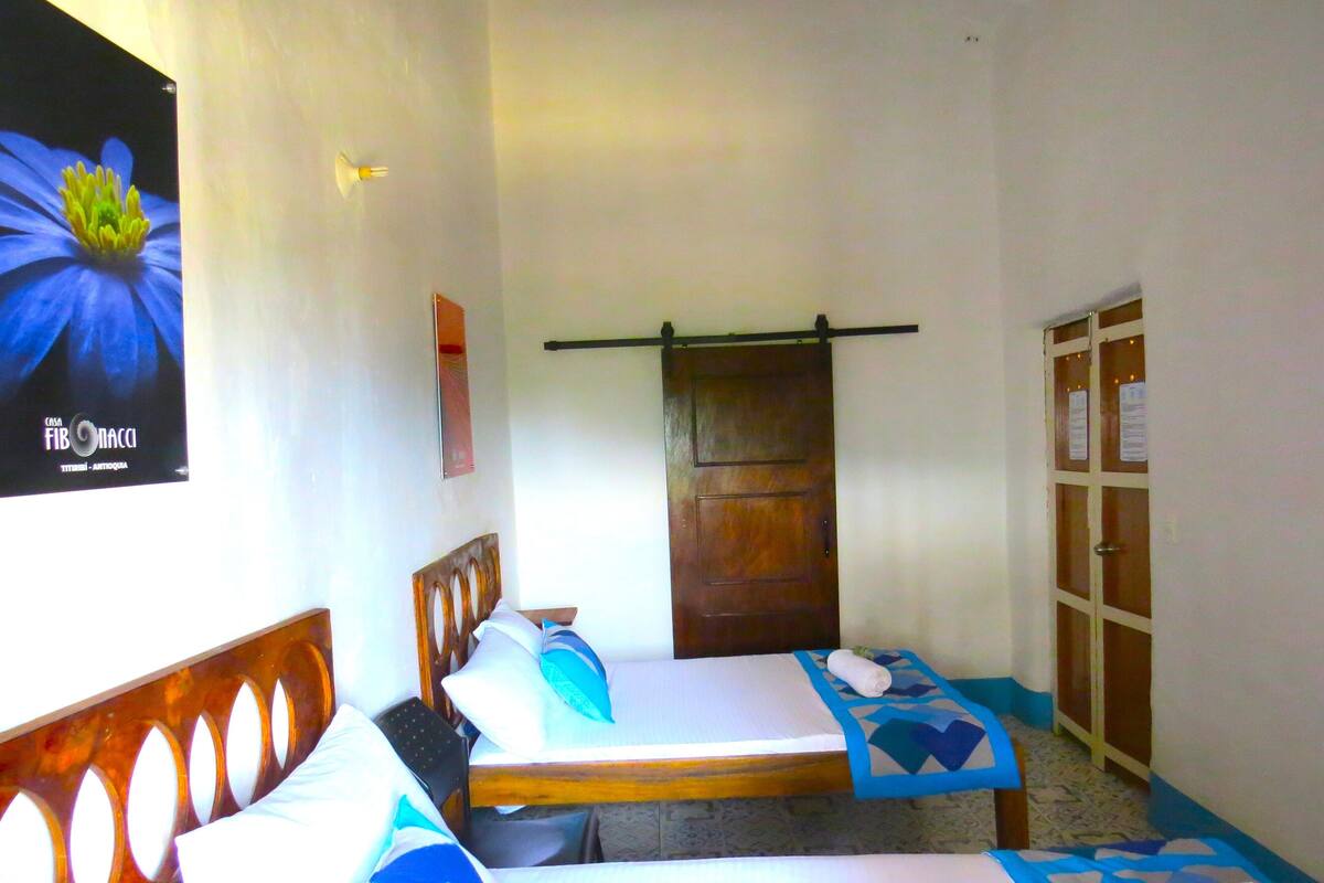 A hotel room with two single beds, a wooden door, a small table, and a wall-mounted light fixture.