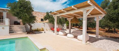 A pool area with a pergola, outdoor seating, and a stone building in the background.