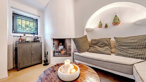 A cozy living room with a wooden coffee table, a sofa with patterned cushions, a fireplace, and a window with a view of trees. - Casale di Amelì 5, Emma Villas (Ostuni)