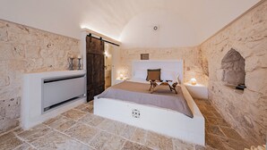 A bedroom with stone walls, a bed with a headboard, bedside tables with lamps, and a wooden door.