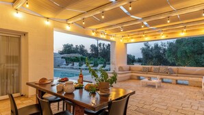 A covered outdoor dining area with a table set for four, a sofa, and a pool in the background.