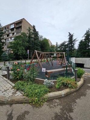A playground with a swing set and a fenced area, surrounded by greenery and a building in the background. - Tbilisi Gardens One Bedroom With Pool (Tbilisi)