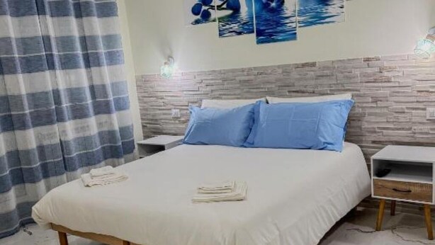 A neatly made bed with white bedding and blue pillows, a wooden headboard, a white nightstand with a drawer, and a striped curtain.