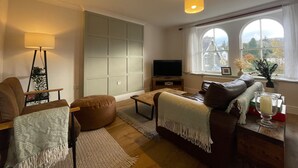 Un salon avec un canapé, un fauteuil et un téléviseur posé sur un meuble. Une fenêtre offre une vue sur des immeubles et des arbres.