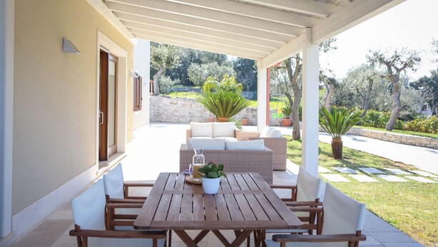 A covered patio with a wooden table and chairs, a wicker sofa, and potted plants.