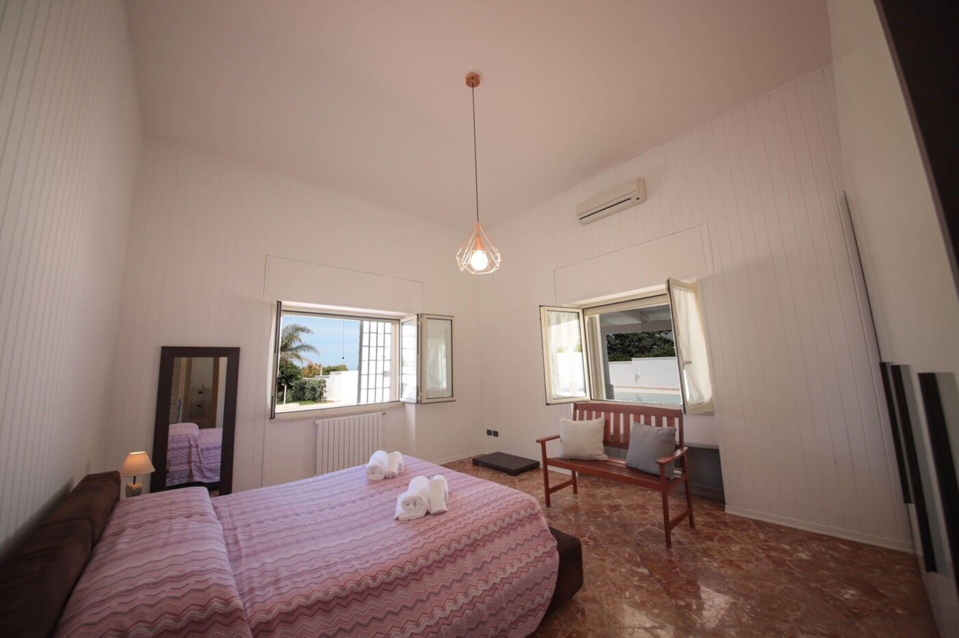 Une chambre à coucher avec un lit, deux fenêtres, un radiateur, un miroir et un luminaire suspendu.