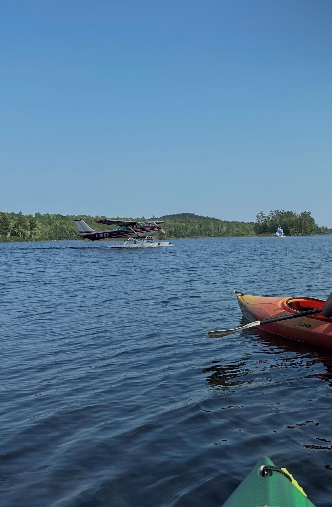 En stilla sjö med ett sjöflygplan på vattnet, en segelbåt i fjärran och kajaker längs stranden.