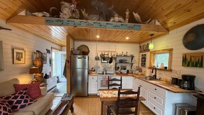 Interior acogedor de una cabaña con techo de madera, un comedor, una cocina con refrigerador y una sala de estar con sofá y libros.