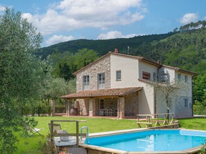 Une maison à deux étages avec une façade en pierre, une piscine et une vue sur une colline boisée.