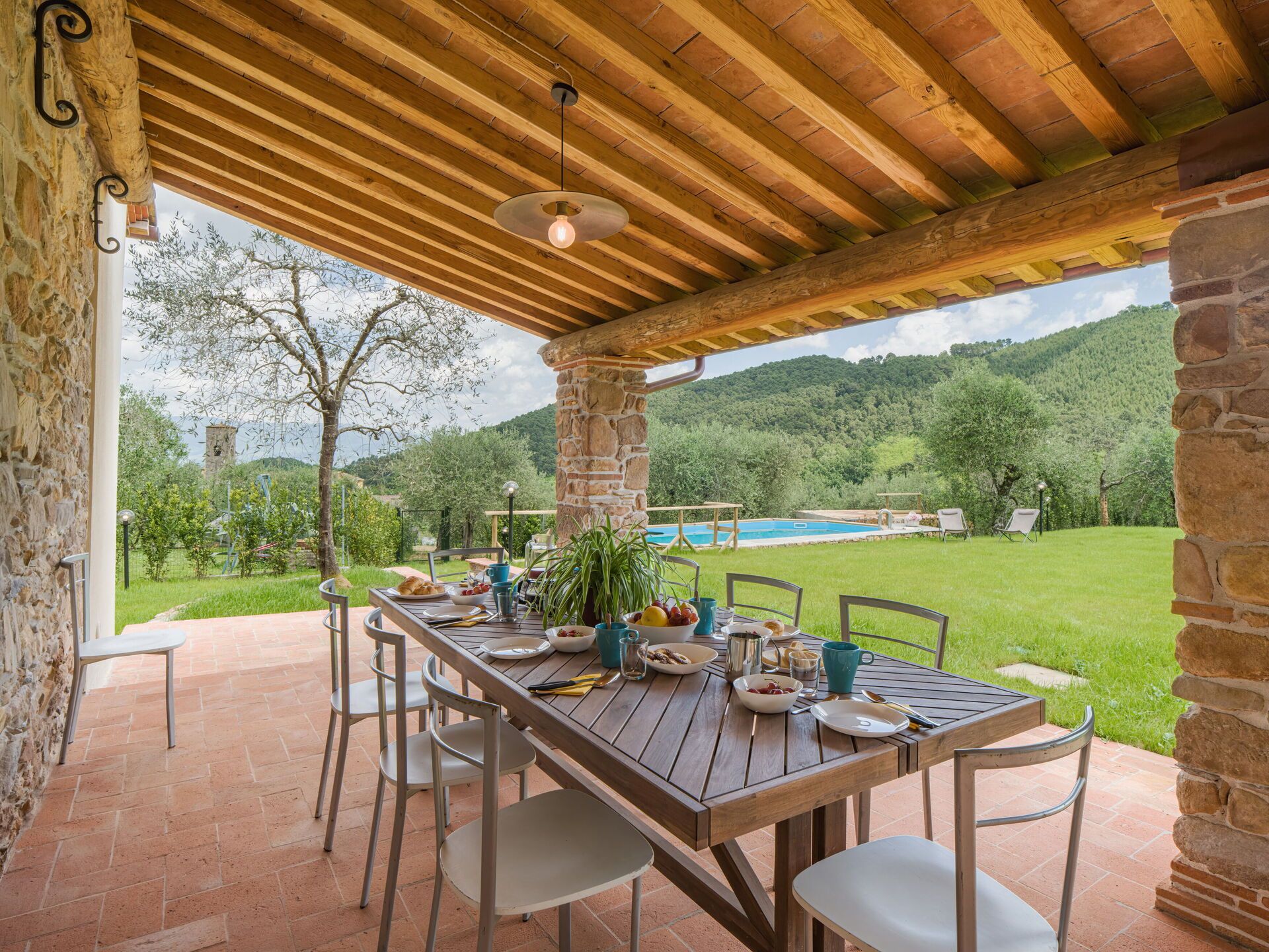 An outdoor dining area with a wooden table and chairs, a stone pillar, and a view of a swimming pool and greenery.