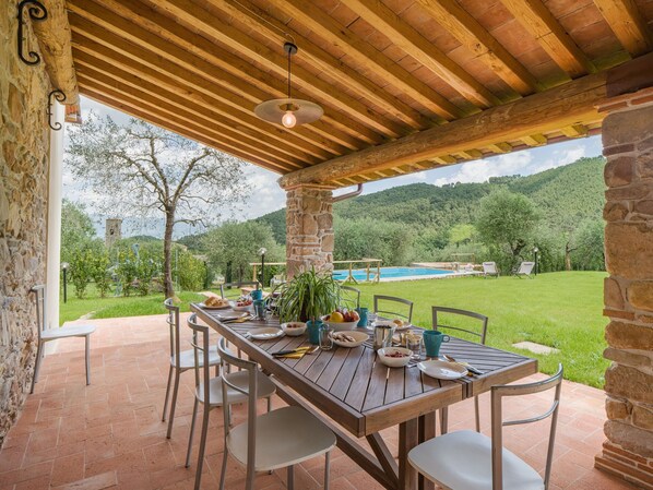 Un espace de restauration en plein air avec une table et des chaises en bois, un pilier en pierre et une vue sur une piscine et la verdure.