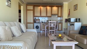 Una sala de estar con sofá, una mesa de centro y un comedor con sillas y una mesa. Se ve una cocina al fondo con armarios y electrodomésticos.