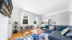 Una sala moderna con un sofá azul oscuro, una mesa de centro de madera y un tapete de diseño azul. Se observa un comedor con mesa y sillas, y una cocina con armarios blancos.