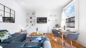 Una sala de estar moderna con un sofá gris, una mesa de centro y un comedor con mesa y sillas. Hay una estantería con objetos decorativos y un ventanal con vistas a edificios.