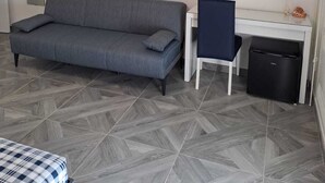 Une pièce avec un canapé gris, un bureau avec une chaise, une poubelle noire et un téléviseur fixé au mur.