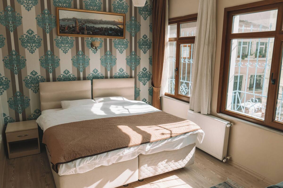 A bedroom with a bed, a nightstand, a window with a view of buildings, and patterned wallpaper.