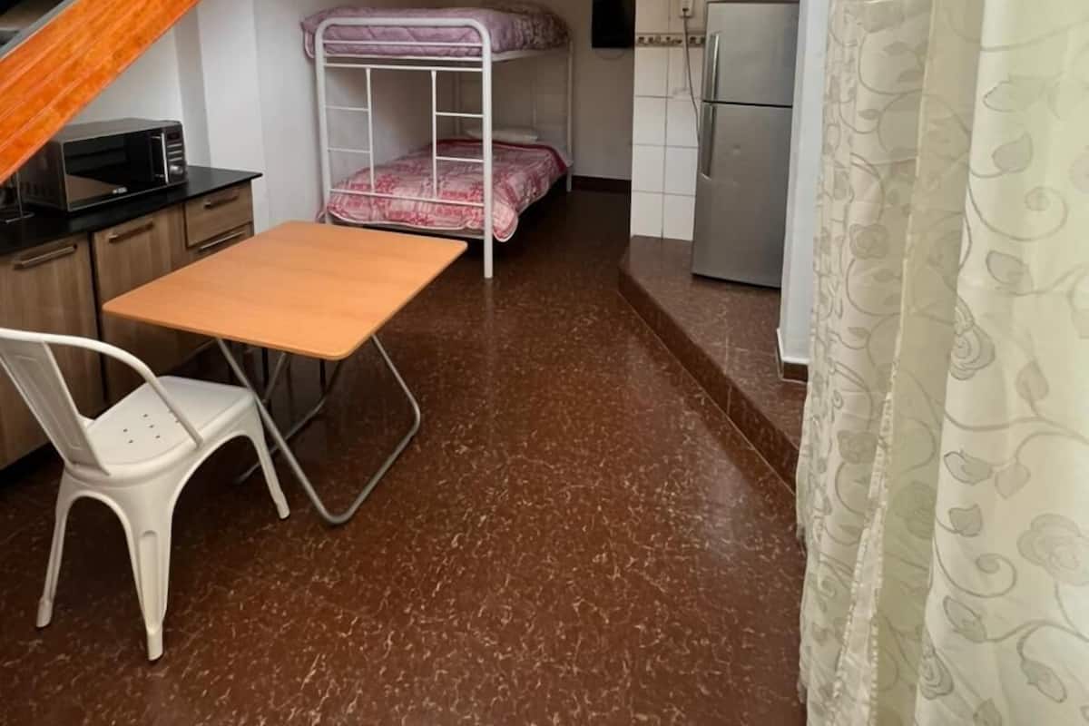 Une chambre d’étudiant de taille réduite, équipée de lits superposés, d’un coin cuisine, d’une petite table et d’un escalier.