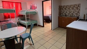 A dormitory room with bunk beds, a small kitchen area, and a dining table with chairs.