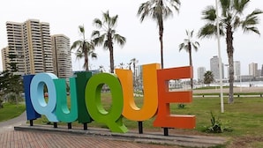 A colorful sign spelling "QUIQUE" on a wooden boardwalk with palm trees and buildings in the background.