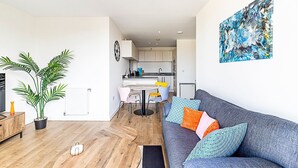 En moderne stue med en grå sofa, farverige puder, et spiseområde med et hvidt bord og stole, og et køkken i baggrunden.