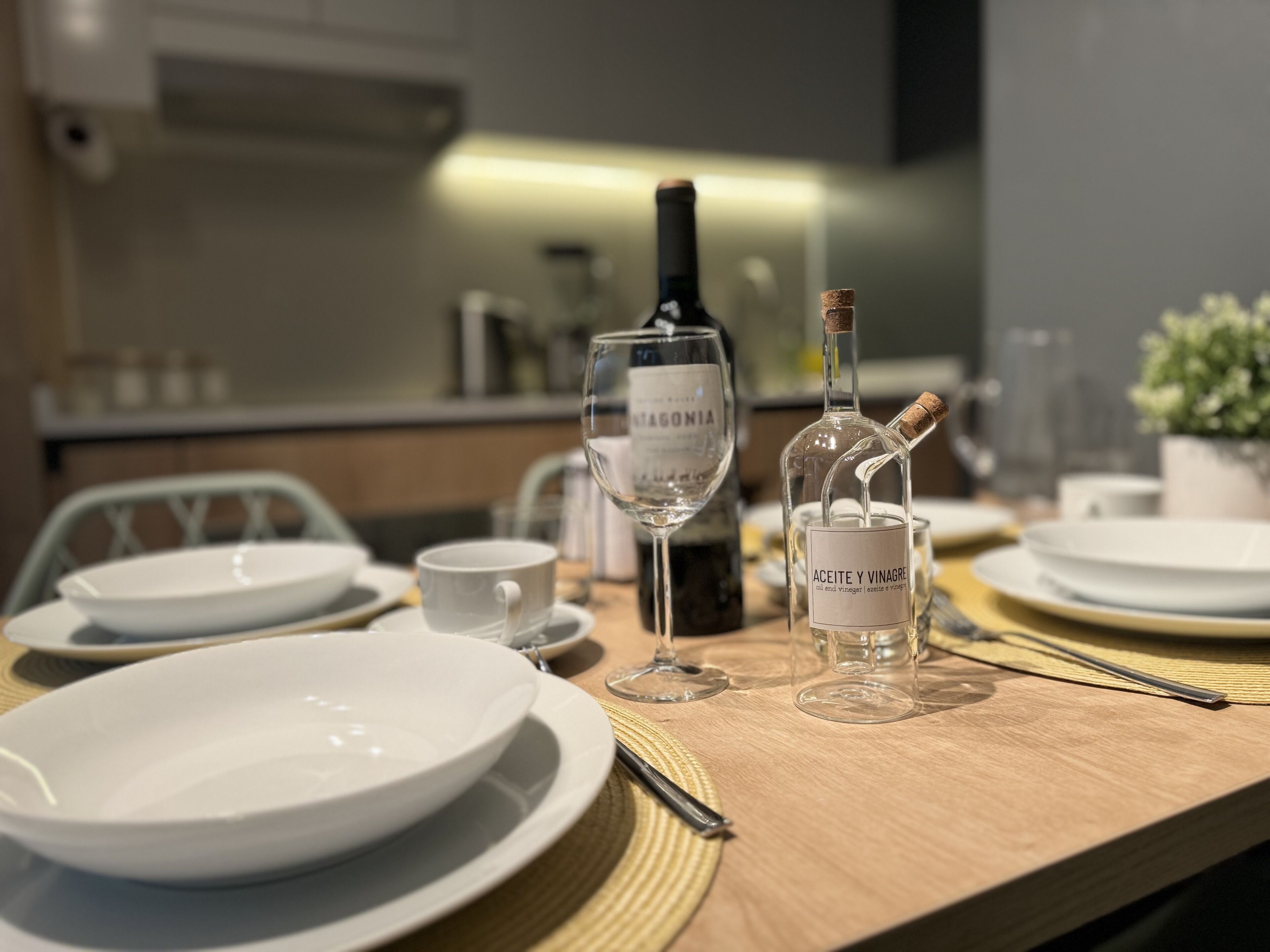 A dining table set with white plates, bowls, and wine glasses, with a bottle of wine and a bottle of vinegar labeled "Aceite y Vinagre".
