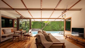 A modern living room with a wooden floor, a pool, and large windows overlooking a lush garden. - Private villa only a short stroll to the beach/Organic shop (Cobano)