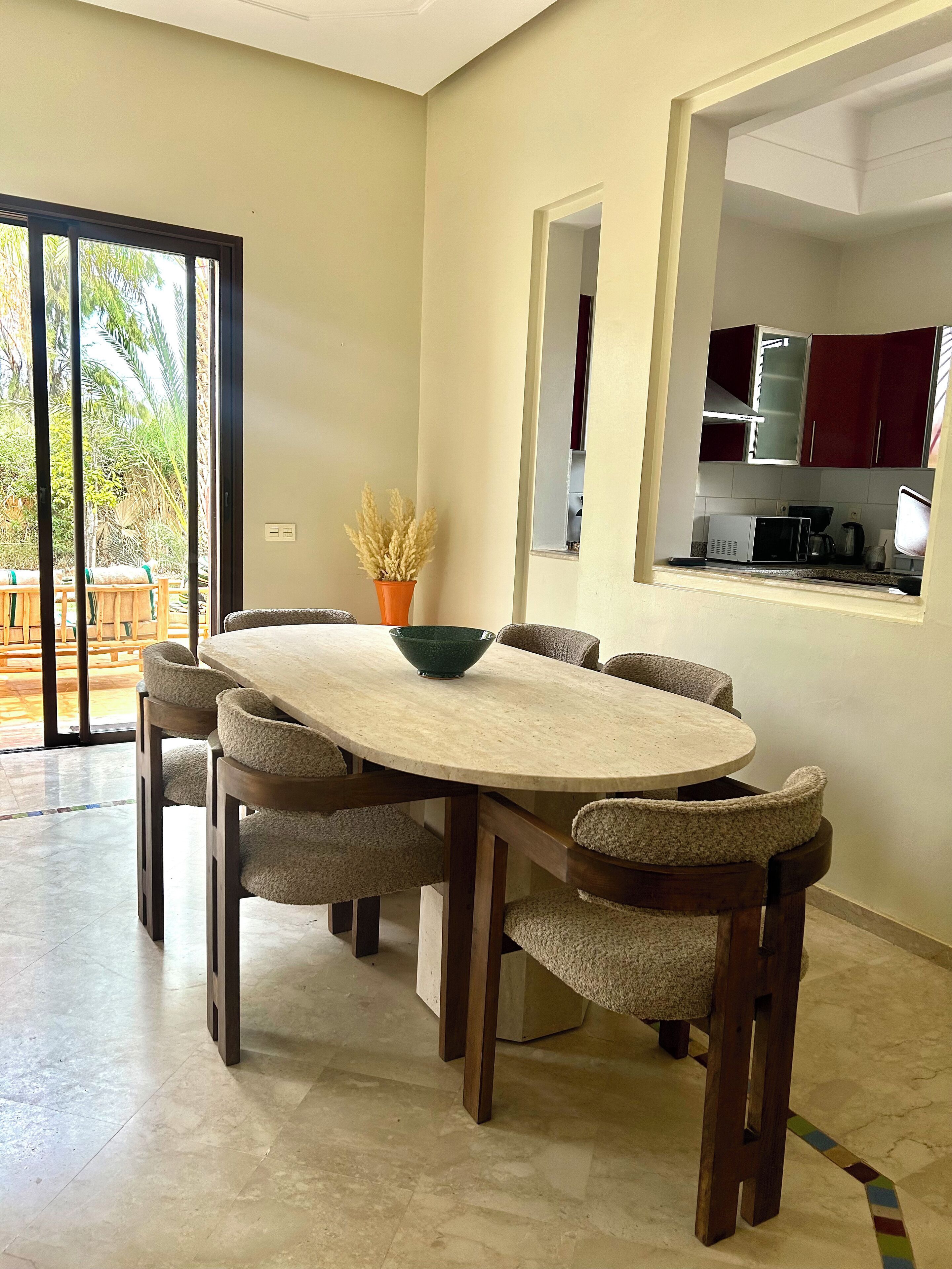 Un coin repas avec une table ronde, six chaises et un saladier. Une porte-fenêtre coulissante donne sur un espace extérieur aménagé avec une table et des chaises en bois. La cuisine est visible à travers une arche, avec des meubles de couleur rouge.