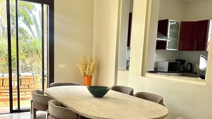 A dining area with a round table, six chairs, and a bowl. There is a sliding glass door leading to an outdoor area with a wooden table and chairs. The kitchen is visible through an archway with red cabinets. - Fabulous family villa in Marrakech (Marrakech)