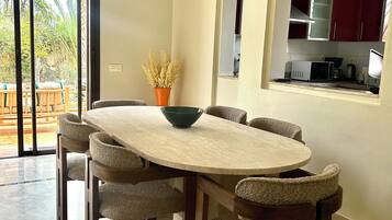 Un coin repas avec une table ronde, six chaises et un saladier. Une porte-fenêtre coulissante donne sur un espace extérieur aménagé avec une table et des chaises en bois. La cuisine est visible à travers une arche, avec des meubles de couleur rouge.