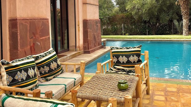 A poolside area with wooden furniture and cushions, a pool, and a building with a beige facade. - Fabulous family villa in Marrakech (Marrakech)