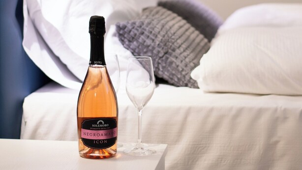 A bed with a blue headboard, white pillows, and a grey blanket. A bottle of wine and a glass are placed on a bedside table.