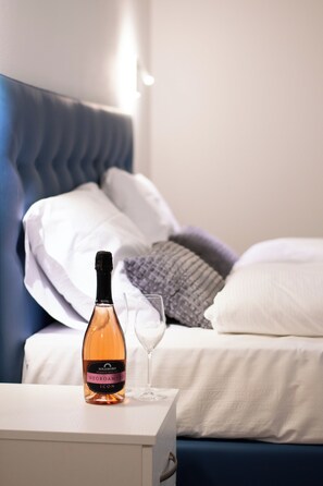 A bed with a blue headboard, white pillows, and a grey blanket. A bottle of wine and a glass are placed on a bedside table.