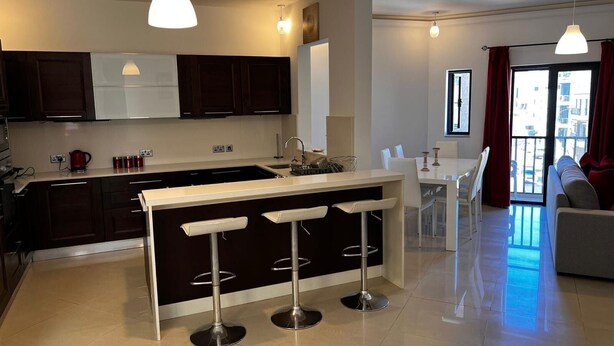 A modern kitchen with dark cabinets, a white countertop, and a central island with bar stools. There is a dining area with a white table and chairs, and a living area with a sofa and a large window. - Naxxar 3-Bedroom Spacious Family Home with Private Roof by Malta Weekend (Naxxar)