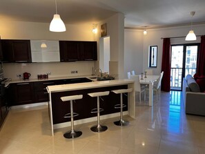 A modern kitchen with dark cabinets, a white countertop, and a central island with bar stools. There is a dining area with a white table and chairs, and a living area with a sofa and a large window.
