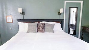 A neatly made bed with white linens and three pillows, a dark wooden headboard, two wall-mounted lamps, and a framed picture on the wall.