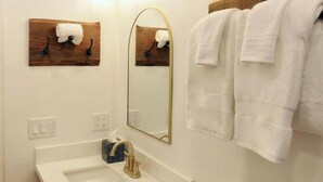 Un baño con lavamanos blanco, grifo dorado, espejo y dos ganchos para toallas con toallas blancas.