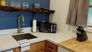 Una cocina compacta con un lavamanos, un microondas, un juego de cuchillos y una estantería con tazas y vasos.