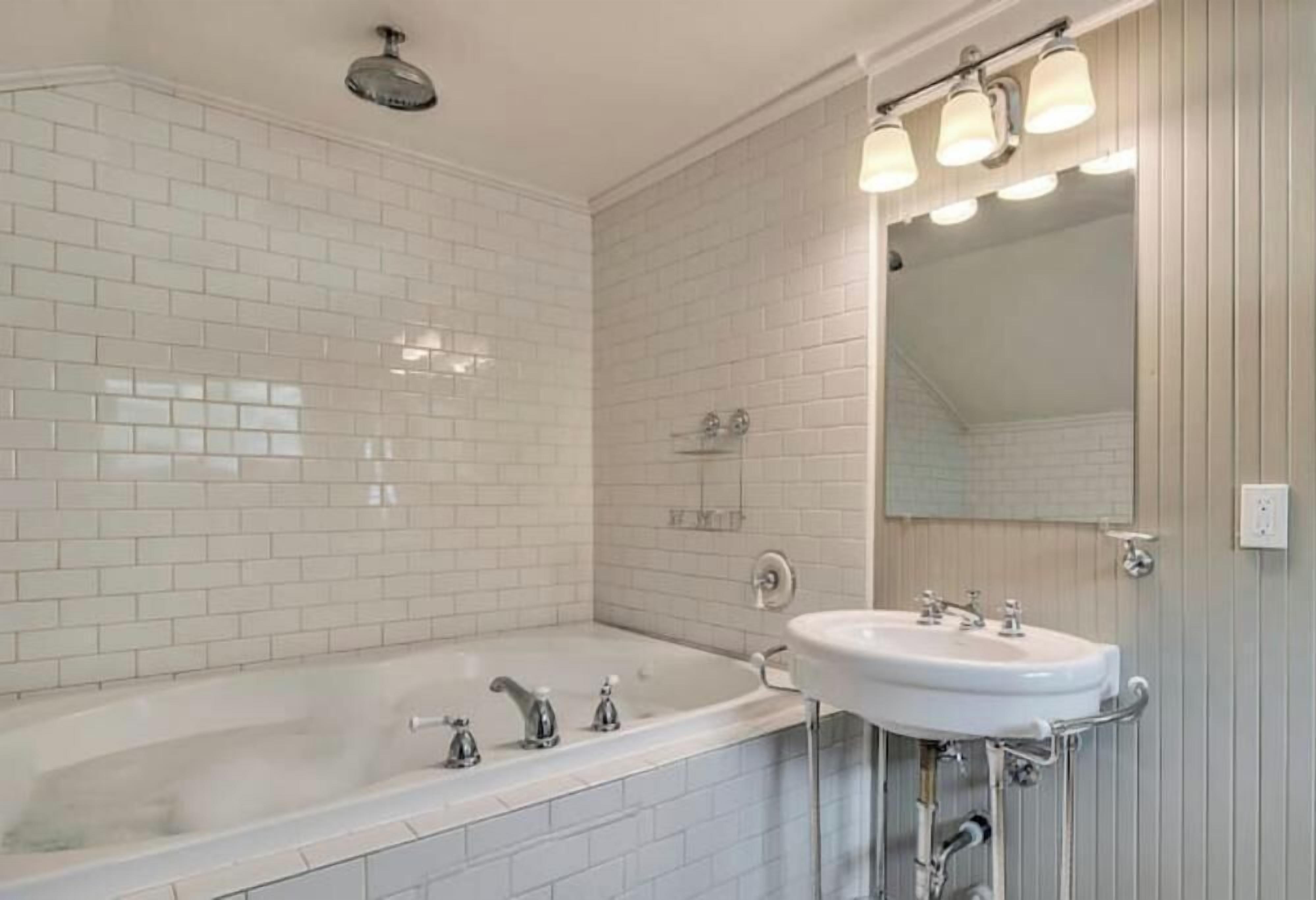 A bathroom with a white-tiled wall, a bathtub, a sink, and a mirror with lights.