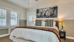 A bedroom with a bed, a ceiling fan, a wall-mounted artwork of barrels, and a small table with a lamp and flowers.