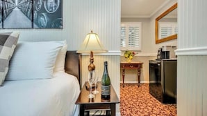 A hotel room with a bed, a bedside table with a lamp, a bottle of wine, and a glass. There is a small table with a vase of flowers and a coffee machine. - Luxury King Room. Steps to Napa Valley Wineries, Spas & Fine Dining! (Calistoga)
