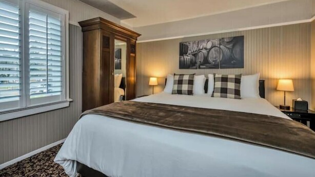 Une chambre d’hôtel avec un grand lit, deux lampes de chevet, une fenêtre avec des stores, une tête de lit en bois et une œuvre d’art accrochée au mur.
