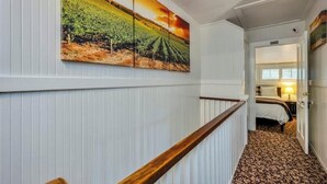 Un couloir d’hôtel avec une rambarde en bois, une grande peinture représentant un vignoble accrochée au mur, et une chambre avec un lit et une table de chevet.