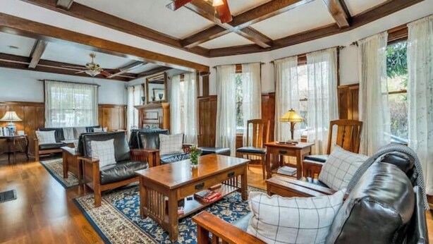 Ein geräumiges Wohnzimmer mit Holzmöbeln, einem Deckenventilator und großen Fenstern mit Vorhängen.