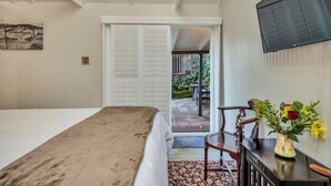 A hotel room with a bed, a desk with a vase of flowers, a television on the wall, and a view of the outdoor patio. - Charming King Room, Private Entrance and Garden Area. Pet-Friendly! (Calistoga)