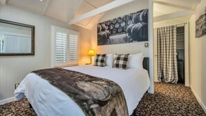 A bedroom with a large bed, a headboard with a barrel pattern, a window with blinds, a lamp, and a checkered curtain. - Charming King Room, Private Entrance and Garden Area. Pet-Friendly! (Calistoga)